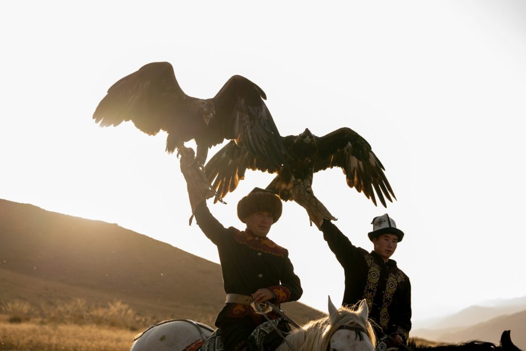 Two men on horseback holding eagles at sunset.